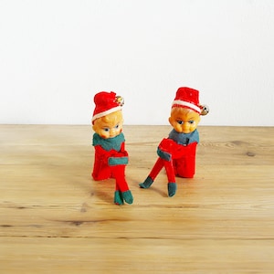 Könnte beinhalten: Zwei rote Weihnachtswichtel aus Filz mit grünem und weißem Besatz und Glöckchen an ihren Mützen. Die Wichtel sitzen auf einer Holzoberfläche.