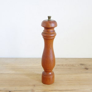 May include: A wooden pepper mill with a traditional design. The mill is made of warm-toned wood and has a brass-coloured top. It has a rounded top and a slender, curved body, standing on a wooden surface.