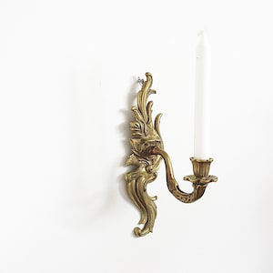 May include: Ornate brass wall sconce with a white candle. The sconce features a detailed floral design and a curved arm to hold the candle. A classic decorative piece for home decor.