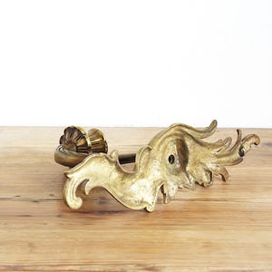 May include: An antique brass-colored decorative door handle with ornate, swirling leaf-like details. The handle features a sunburst-style design on one end and a flowing, organic shape on the other. It rests on a wooden surface.