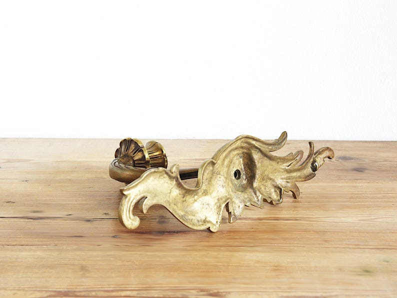 May include: An antique brass-colored decorative door handle with ornate, swirling leaf-like details. The handle features a sunburst-style design on one end and a flowing, organic shape on the other. It rests on a wooden surface.