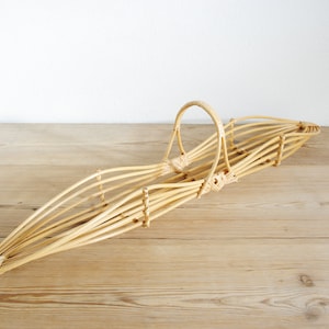 Pode incluir: Uma cesta de vime tecida com uma alça, feita de vime natural de cor castanho claro. A cesta é longa e estreita, com uma forma ligeiramente curva.