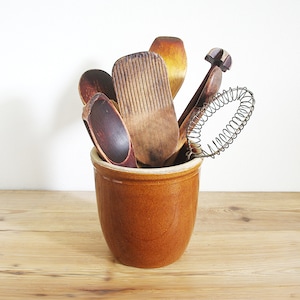 May include: A collection of wooden kitchen utensils, including spoons, a butter paddle, and a whisk, are displayed in a brown ceramic crock. The utensils are various shades of brown and tan, and the crock sits on a light wood surface.
