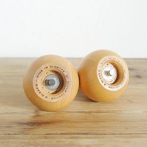 May include: Two wooden salt and pepper shakers with a light brown finish. The shakers have a round shape and are made of wood. The text "MADE IN DENMARK BY JUELSSEN" is printed on the bottom of each shaker.