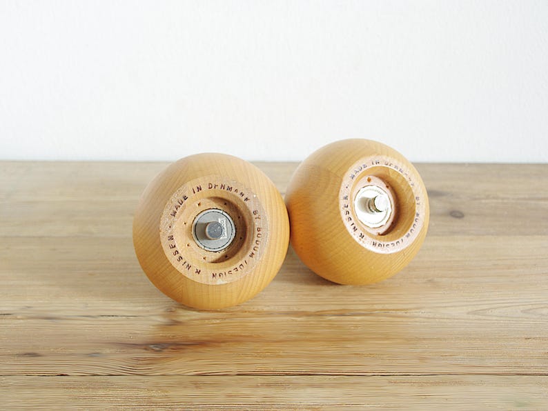 May include: Two wooden salt and pepper shakers with a light brown finish. The shakers have a round shape and are made of wood. The text "MADE IN DENMARK BY JUELSSEN" is printed on the bottom of each shaker.
