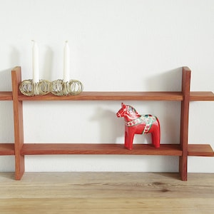 May include: A two-tiered wooden shelf with a red Dala horse figurine and three white candles in gold holders. The shelf is made of reddish-brown wood and is mounted on a white wall.