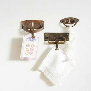 Può includere: Tre maniglie per porte in ottone antico montate su una parete bianca. Le maniglie sono di stili diversi, una è un semicerchio, un'altra è un rettangolo e l'ultima è una maniglia curva con un buco della serratura. Un fazzoletto bianco con bordo di pizzo è appeso alla maniglia semicircolare.