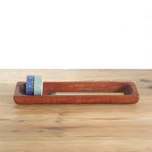 Op de afbeelding: Een rechthoekig, roodbruin houten dienblad met twee kleine rollen washi-tape met patronen en een enkel potlood. Het dienblad heeft een gladde, uitgesneden binnenkant en afgeronde randen voor het opbergen van kleine bureauartikelen.