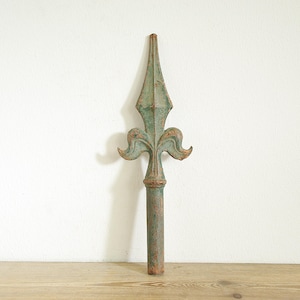 May include: A green metal decorative finial with a fleur-de-lis design. The finial is weathered and has a rustic look.