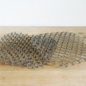 May include: A piece of chainmail armour, made from interconnected metal rings. The chainmail has a dull silver hue and rests on a light brown wooden surface. The texture of the chainmail is visible, showing the individual rings.