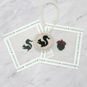 May include: A beige and green squirrel-themed needlepoint ornament with a matching acorn design. The ornament is round with a cream-colored border and a cream-colored cord. Two white square needlepoint canvases with a squirrel and acorn design are also present. The text "WINSTON TALES" is printed on the canvases.