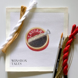 May include: A needlepoint canvas with a red and gold caviar tin design. The canvas is surrounded by embroidery floss in white, tan, gold, and red. The canvas also includes the text "Winston Tales".