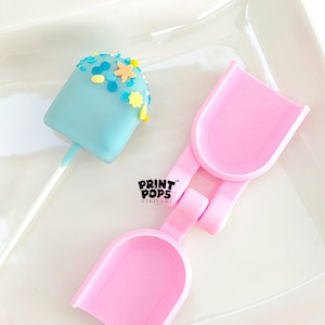 May include: A light blue cake pop with colorful sprinkles and a pink shovel-shaped tool. The cake pop is decorated with blue, yellow, and orange sprinkles. The pink tool has the text "PRINT POPS STRIVAMI".