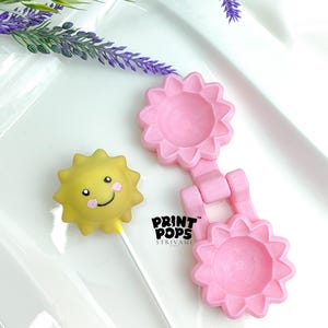 May include: A yellow sun-shaped cake pop with a smiling face and a pink blush, next to a pink flower-shaped mold. The mold is connected by a hinge. The text "PRINT POPS STRIVAMI" is visible.