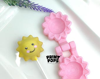 Peut inclure: Un cake pop jaune en forme de soleil avec un visage souriant et des joues roses, à côté d'un moule rose en forme de fleur. Le moule est relié par une charnière. Le texte "PRINT POPS STRIVAMI" est visible.