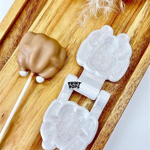 May include: A light brown cake pop shaped like a fist on a white stick, next to a white plastic mold with the text "PRINT POPS". The items are on a wooden tray.
