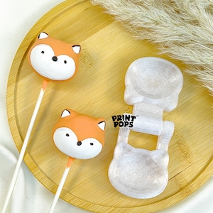 K&ouml;nnte beinhalten: Zwei orange-wei&szlig;e, fuchsf&ouml;rmige Cake Pops mit schwarzen Augen und Ohren auf wei&szlig;en Stielen liegen auf einem Holztablett. Neben den Cake Pops befindet sich eine transparente, katzenf&ouml;rmige Form. Der Text "PRINT POPS" ist sichtbar.