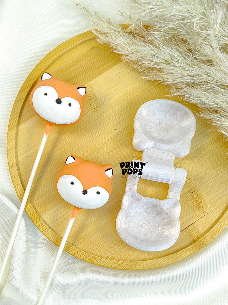 K&ouml;nnte beinhalten: Zwei orange-wei&szlig;e, fuchsf&ouml;rmige Cake Pops mit schwarzen Augen und Ohren auf wei&szlig;en Stielen liegen auf einem Holztablett. Neben den Cake Pops befindet sich eine transparente, katzenf&ouml;rmige Form. Der Text "PRINT POPS" ist sichtbar.