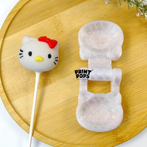 K&ouml;nnte beinhalten: Ein wei&szlig;er Hello Kitty Cake Pop mit roter Schleife und eine transparente Kunststoffform in Form von Hello Kitty. Die Form tr&auml;gt den Text "PRINT POPS". Die Artikel befinden sich auf einem Holztablett.