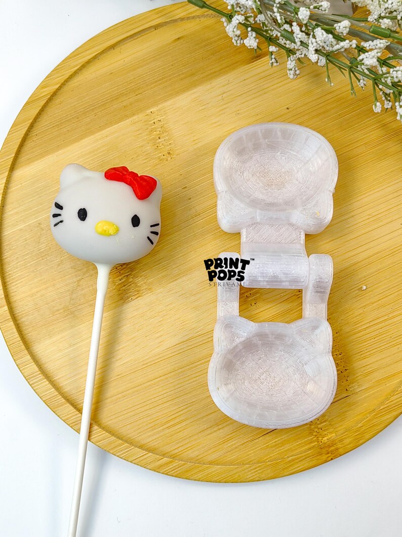 K&ouml;nnte beinhalten: Ein wei&szlig;er Hello Kitty Cake Pop mit roter Schleife und eine transparente Kunststoffform in Form von Hello Kitty. Die Form tr&auml;gt den Text "PRINT POPS". Die Artikel befinden sich auf einem Holztablett.