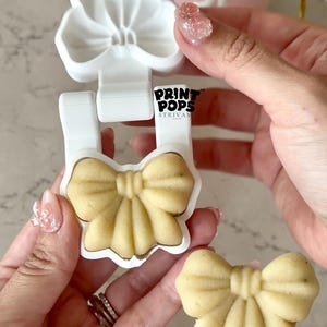 May include: A white plastic bow-shaped mold with the text PRINT POPS STRIVAN printed on the center. The mold is open, revealing a beige bow-shaped treat inside, while a finished beige bow-shaped treat on a clear stick is held below it.
