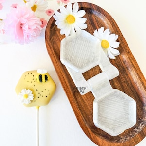 May include: A wooden tray holds two clear, hexagonal molds and a bee-themed cake pop. The cake pop is yellow with a black and yellow bee and a white daisy. Artificial flowers and a floral patterned cloth are in the background.
