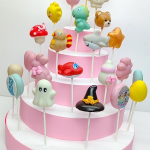 K&ouml;nnte beinhalten: Ein mehrst&ouml;ckiger rosa Kuchenst&auml;nder mit zahlreichen Cake Pops. Die Cake Pops sind als Tiere, Objekte und Charaktere gestaltet, darunter ein Hai, ein B&auml;r und ein Hexenhut. Der Kuchenst&auml;nder tr&auml;gt das Wort "STRIVAMI" und ein "PRINT POPS"-Schild.