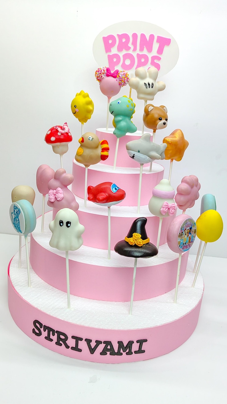 K&ouml;nnte beinhalten: Ein mehrst&ouml;ckiger rosa Kuchenst&auml;nder mit zahlreichen Cake Pops. Die Cake Pops sind als Tiere, Objekte und Charaktere gestaltet, darunter ein Hai, ein B&auml;r und ein Hexenhut. Der Kuchenst&auml;nder tr&auml;gt das Wort "STRIVAMI" und ein "PRINT POPS"-Schild.