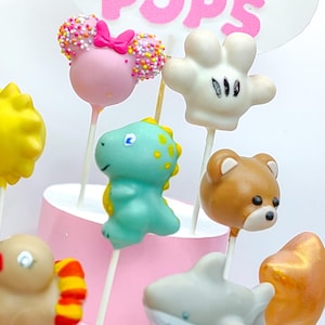 K&ouml;nnte beinhalten: Eine Sammlung bunter Cake Pops am Stiel, darunter ein rosa Cake Pop im Minnie-Maus-Stil, ein gr&uuml;ner Dinosaurier, ein brauner B&auml;r und ein wei&szlig;er Cake Pop in Mickey-Maus-Form. Die Cake Pops sind auf einem rosa Kuchenst&auml;nder angeordnet, mit der Aufschrift "PRINT POPS".