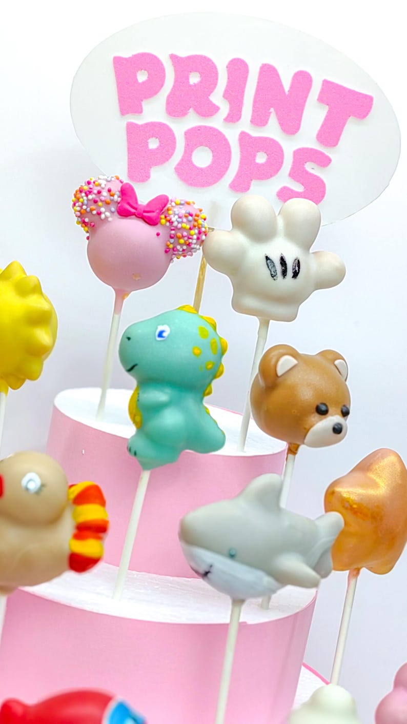K&ouml;nnte beinhalten: Eine Sammlung bunter Cake Pops am Stiel, darunter ein rosa Cake Pop im Minnie-Maus-Stil, ein gr&uuml;ner Dinosaurier, ein brauner B&auml;r und ein wei&szlig;er Cake Pop in Mickey-Maus-Form. Die Cake Pops sind auf einem rosa Kuchenst&auml;nder angeordnet, mit der Aufschrift "PRINT POPS".