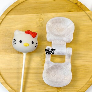 K&ouml;nnte beinhalten: Ein wei&szlig;er Hello Kitty Cake Pop mit roter Schleife und schwarzen Details, neben einer wei&szlig;en Form mit dem Text "PRINT POPS". Die Artikel befinden sich auf einem runden Holztablett.