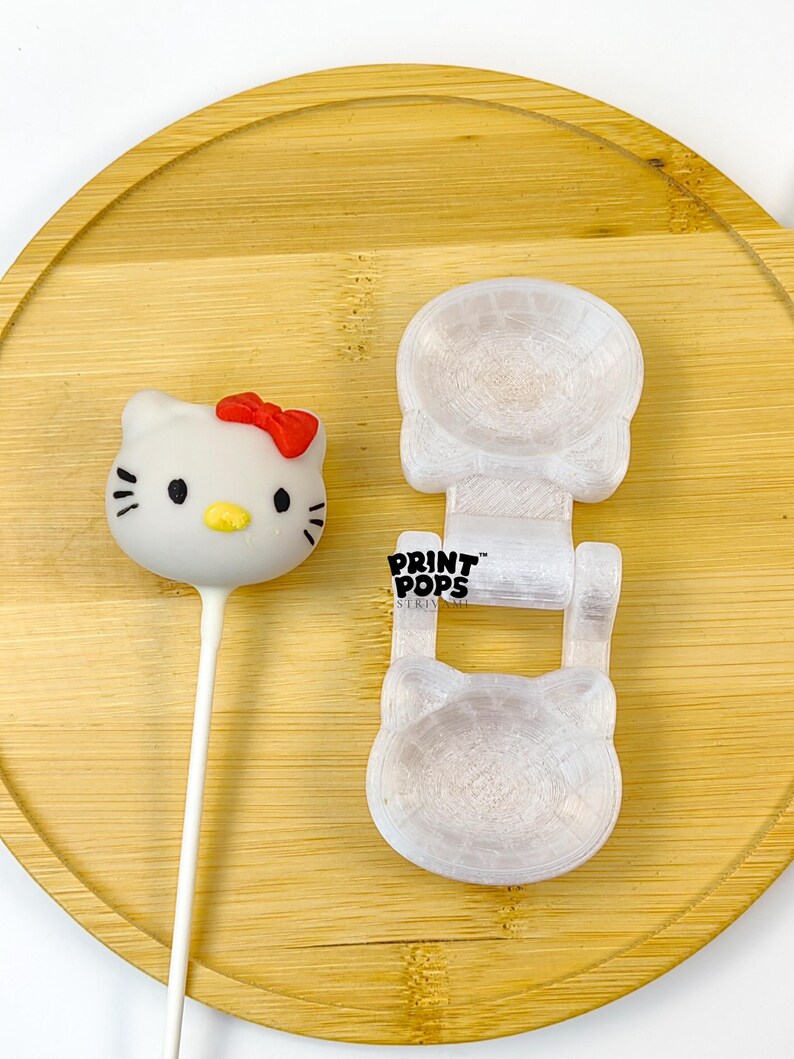 K&ouml;nnte beinhalten: Ein wei&szlig;er Hello Kitty Cake Pop mit roter Schleife und schwarzen Details, neben einer wei&szlig;en Form mit dem Text "PRINT POPS". Die Artikel befinden sich auf einem runden Holztablett.