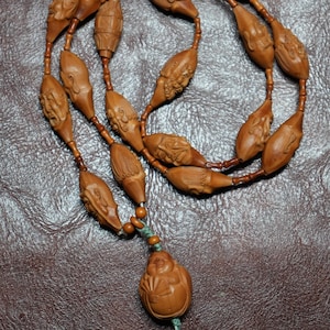 May include: A handcrafted wooden necklace featuring intricately carved beads with human faces and a smiling Buddha pendant. The beads are connected by a brown string, and the pendant is suspended from a green cord. The necklace is a unique piece of jewelry.