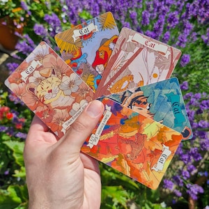 May include: A set of four rectangular trading cards featuring cat illustrations, each labelled with the word Cat. The cards display colourful, detailed artwork of various feline figures amidst floral and botanical patterns, held in a fan shape.