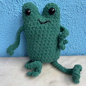 May include: A green crocheted frog plush toy with black eyes and a smiling mouth. The frog is sitting on a white surface.