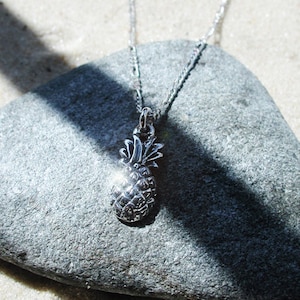 Puede incluir: Un collar con colgante de piña plateada. El colgante es una piña en miniatura detallada, y la cadena es delicada. El collar se muestra sobre una piedra gris, con la luz del sol creando un reflejo brillante en el colgante.