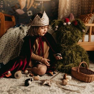 May include: A child wearing a white crown and a black and red cape sits on a patterned rug surrounded by wooden toys, a green furry creature, and a wicker basket. A wooden dollhouse and bed are in the background.