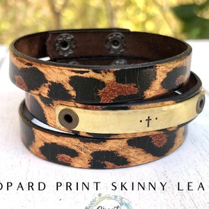May include: A brown leather bracelet with a leopard print design. The bracelet has a gold metal plate with the text ". . . + . . ." engraved on it.
