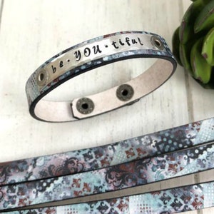 May include: A blue and silver leather bracelet with the words "be you-tiful" engraved on the metal plate. The bracelet has adjustable snaps.