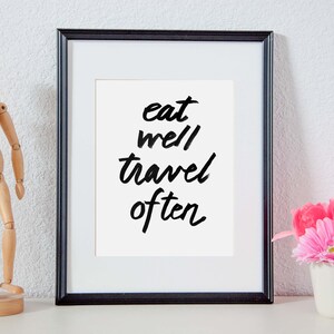 以下が含まれることがあります： 白い背景に白い文字で「eat well travel often」と書かれた黒い額縁のプリント。プリントは白い棚に置かれ、木製の置物と白いマグカップに入ったピンクの花の飾り付けがされている。