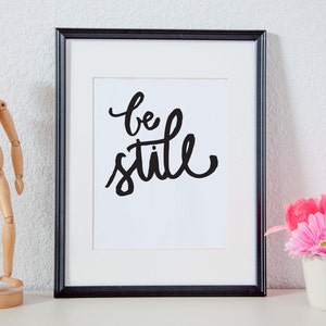 以下が含まれることがあります： 白い背景に白い書体で「be still」と書かれた黒い額縁のプリント。プリントは白い棚に置かれ、木製の置物と白いマグカップに入ったピンクの花の飾り付けがされている。