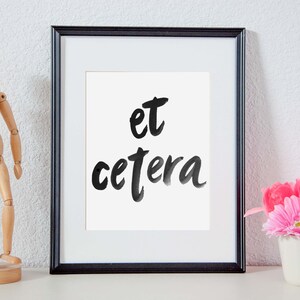 May include: A black framed picture with a white mat and the words "et cetera" written in black ink on a white background. A wooden artist's mannequin stands to the left of the picture and a pink flower arrangement is to the right.