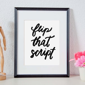 May include: A black framed print with the text "flip that script" in black cursive lettering. The print is on a white background and is sitting on a white shelf with a wooden figurine and a pink flower in a white cup.