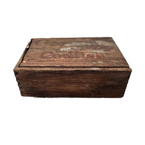 Könnte beinhalten: Eine Vintage-Holzkiste mit dem Wort "Codfish" und anderem Text auf dem Deckel. Die Kiste ist rechteckig und hat ein dunkelbraunes, verwittertes Aussehen, das auf Alter und Gebrauch hindeutet. Die Kiste dient wahrscheinlich zur Aufbewahrung.
