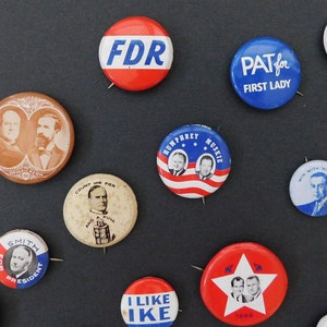May include: A collection of vintage political campaign buttons featuring various candidates and slogans. The buttons are circular and feature a variety of colors, including red, white, blue, and brown. Some of the buttons feature portraits of the candidates, while others feature slogans or logos. The buttons are arranged in a grid pattern on a dark background.