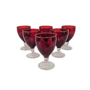 May include: A set of six matching goblets featuring deep red glass bowls and clear, textured glass stems and bases. Each goblet has a rounded bowl shape and a circular foot with a raised, dimpled pattern on the underside of the base.