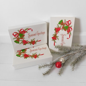 May include: Three white gift boxes with red and green Christmas bells and ribbons. The boxes are stacked on top of each other and say "Season's Greetings" on the front. A small pine branch with red and white ornaments is in the foreground.