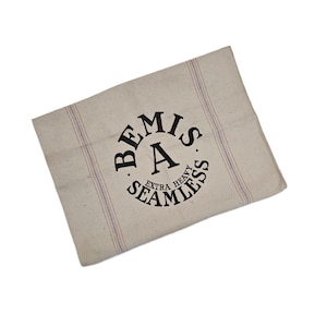 Puede incluir: Tela rectangular beige con rayas azules y rojas y el texto "BEMIS EXTRA HEAVY SEAMLESS" en un diseño circular negro. La tela parece estar hecha de un material natural.