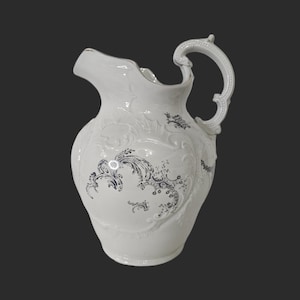 May include: White ceramic jug with a decorative handle and spout. The jug features embossed floral and scrollwork designs with dark blue accents. A classic piece for serving beverages.