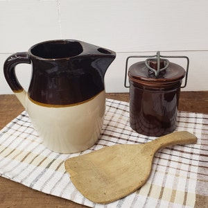 May include: A brown and white ceramic pitcher with a handle, a brown ceramic jar with a wire lid, and a wooden spoon on a checkered tablecloth.